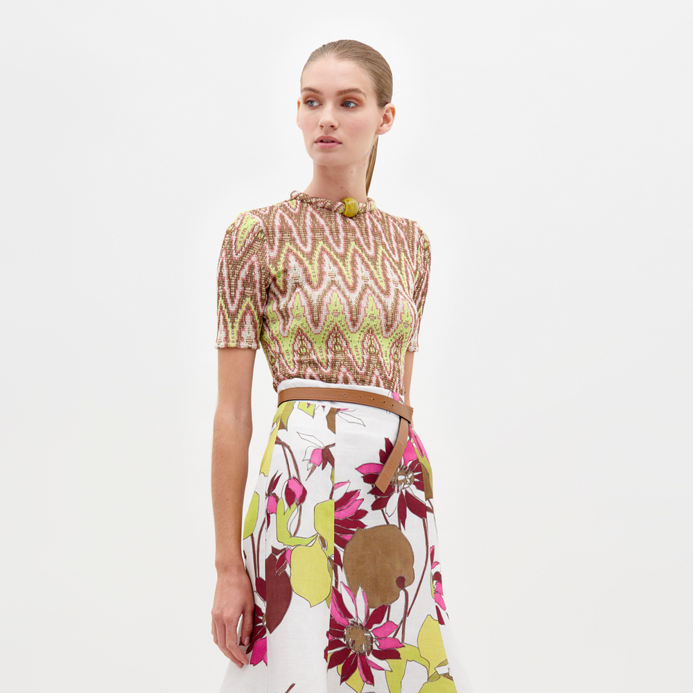 A woman is wearing the Varinia Tshirt Zigzag Neon and a floral skirt, showcasing her fitted silhouette as she looks to her left against a plain background.