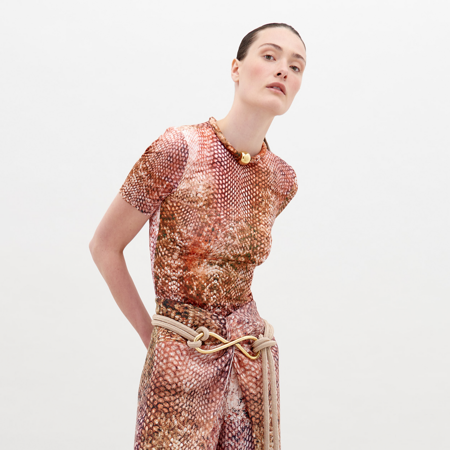 A person poses in a Varinia T-Shirt Peach Animal Print and matching pants, wearing a necklace and belt made of thick rope. Pre-order now to ensure delivery by the ship date, November 15th, 2024.