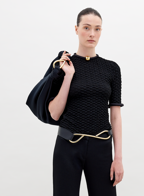 A woman with tied-back hair wears a Venecia Tshirt Black, black pants, and a modern belt, holding a large black bag. She stands against a plain white background, ready to pre-order her outfit by November 15th.