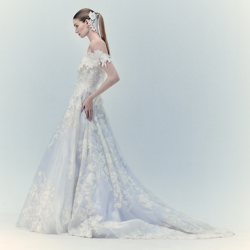 A bride in the exclusive Virginia Dress Pearl, an off-shoulder wedding gown featuring intricate floral details and a long train, stands in profile. Her sleek ponytail is adorned with a white hairpiece. For more details, please inquire within.
