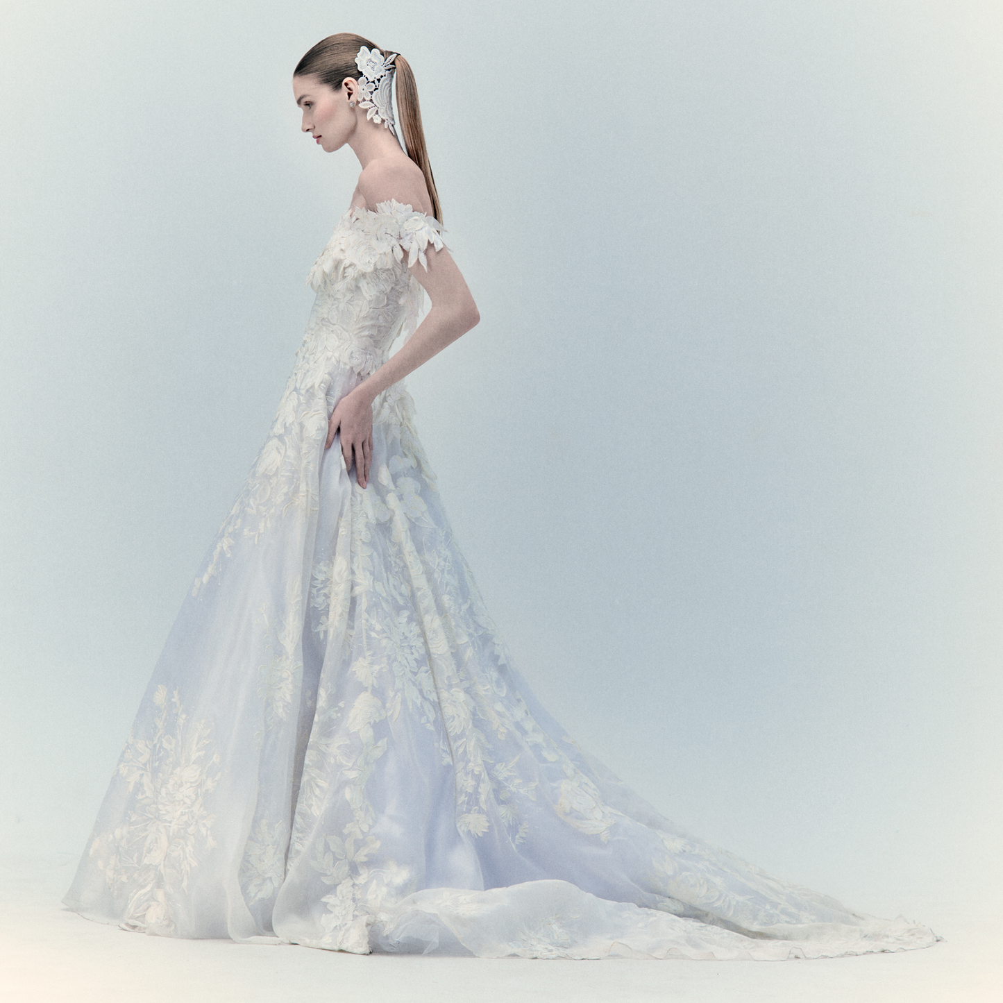 A bride in the exclusive Virginia Dress Pearl, an off-shoulder wedding gown featuring intricate floral details and a long train, stands in profile. Her sleek ponytail is adorned with a white hairpiece. For more details, please inquire within.