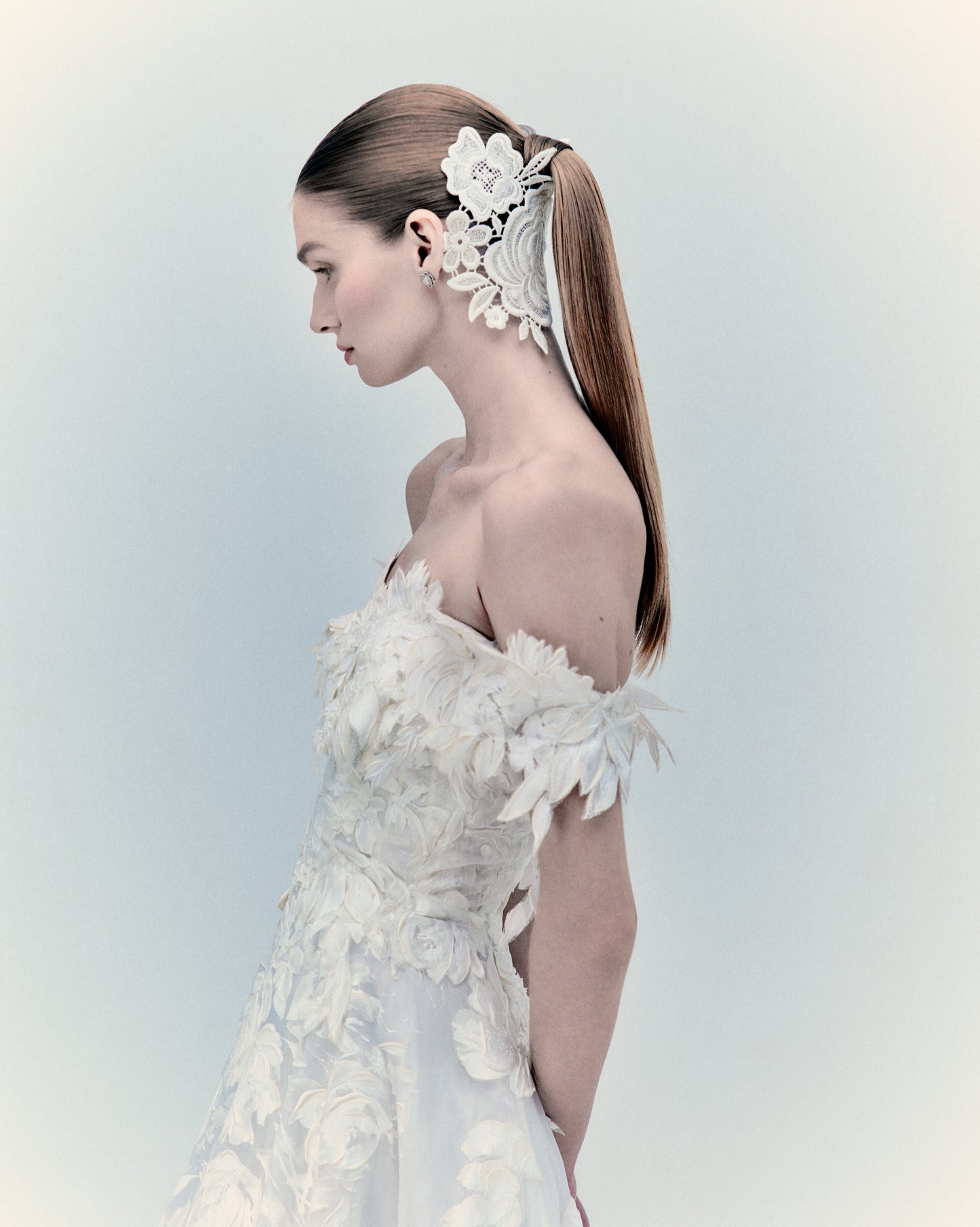 A bride in the exclusive Virginia Dress Pearl, an off-shoulder wedding gown featuring intricate floral details and a long train, stands in profile. Her sleek ponytail is adorned with a white hairpiece. For more details, please inquire within.