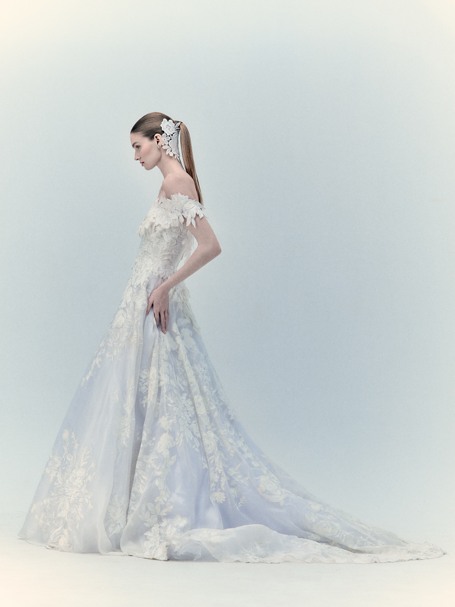 A bride in the exclusive Virginia Dress Pearl, an off-shoulder wedding gown featuring intricate floral details and a long train, stands in profile. Her sleek ponytail is adorned with a white hairpiece. For more details, please inquire within.