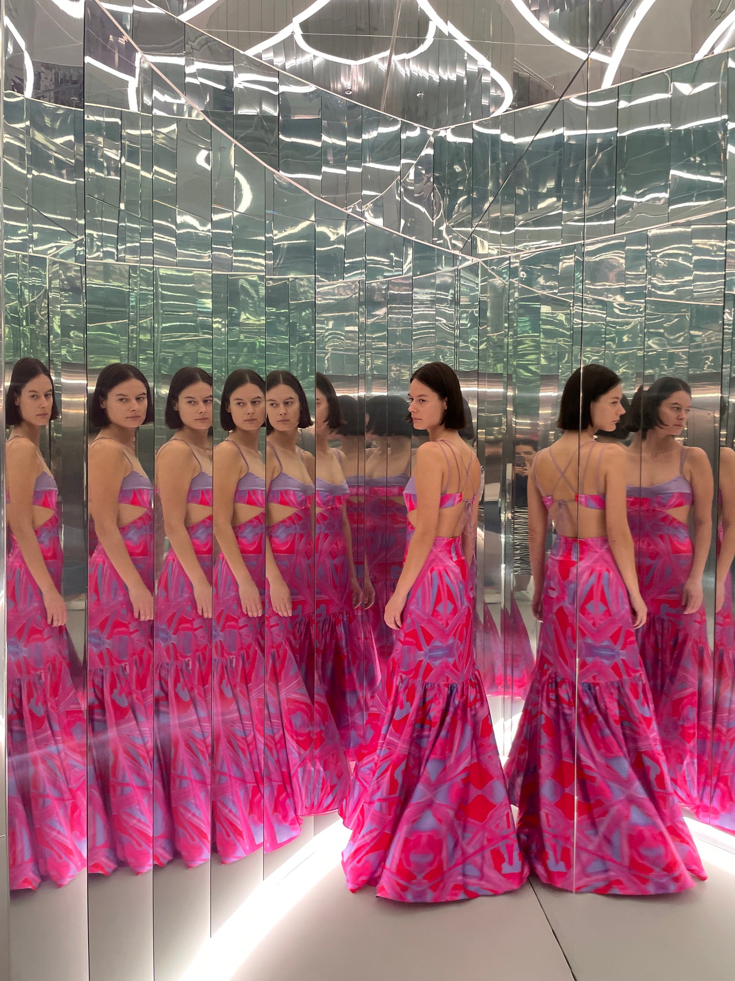 A woman in a pink and purple dress stands in a mirrored room, creating multiple reflections.