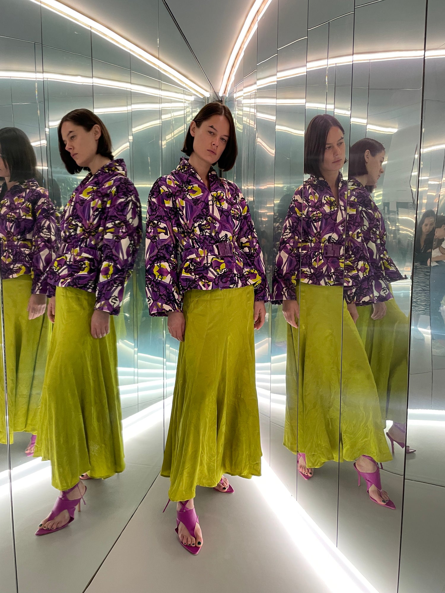 Woman in a purple patterned jacket and green skirt stands in a mirrored room with bright lighting, creating multiple reflections.