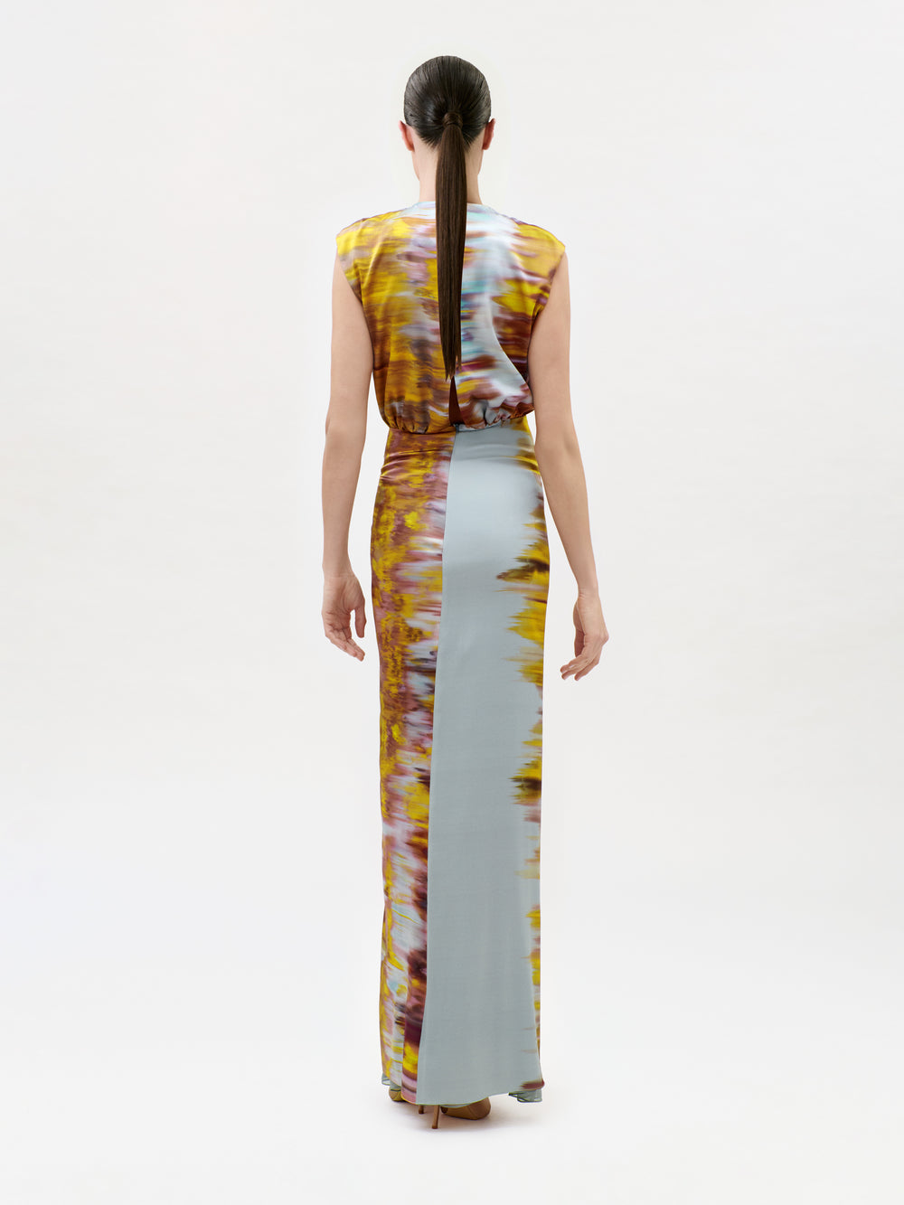 A woman with long brown hair in a ponytail models the Winona Dress, a sleeveless, floor-length gown featuring a yellow, brown, and pale blue tie-dye pattern. She is shown standing with her back to the camera against a white background.