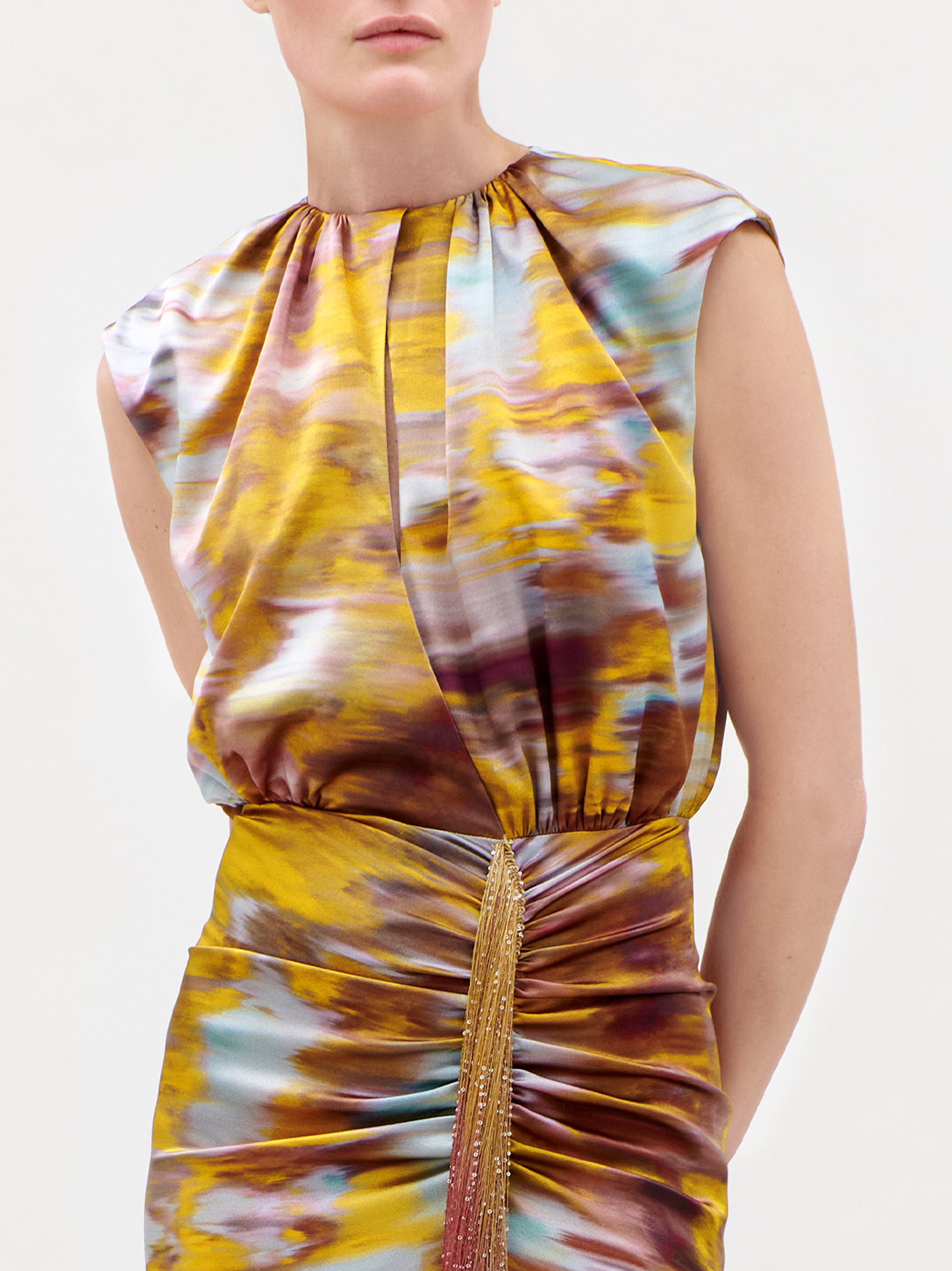 A person wearing the Winona Dress, a sleeveless design featuring a yellow and purple abstract pattern with gathered details at the waist and gold fringe accents. The head is partially out of frame.