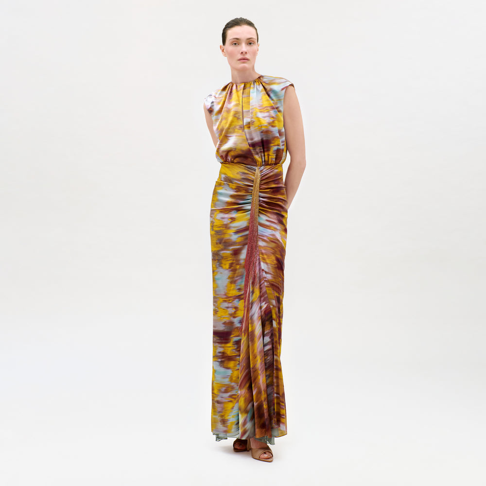 A woman stands against a plain background wearing the Winona Dress, a sleeveless, floor-length dress featuring a colorful tie-dye pattern in yellow, blue, and maroon tones, paired with brown sandals.