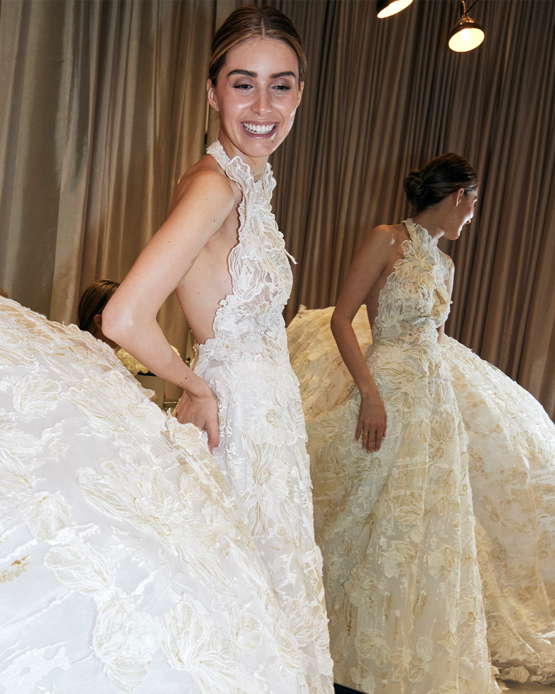 A woman in a sleeveless, intricate lace gown stands in front of a mirror, smiling. The gown features a full skirt with floral patterns and a high-neck design.