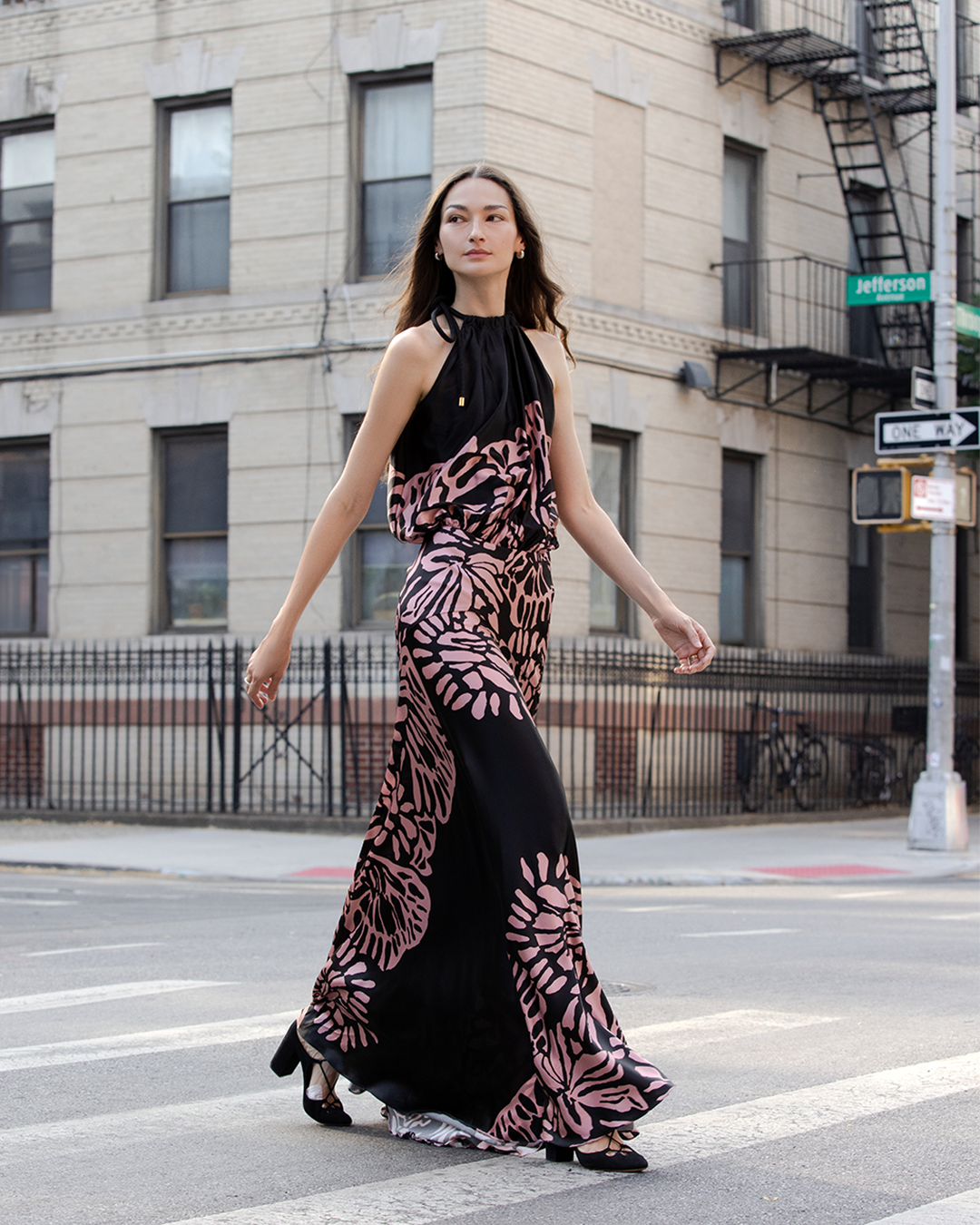 A woman in a black and pink patterned sleeveless dress walks confidently across a city street.