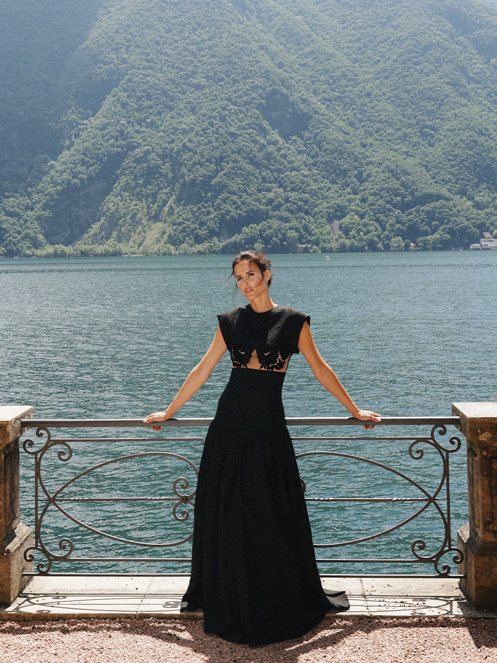 A person in a black dress stands by a railing with a scenic lake and mountains in the background.