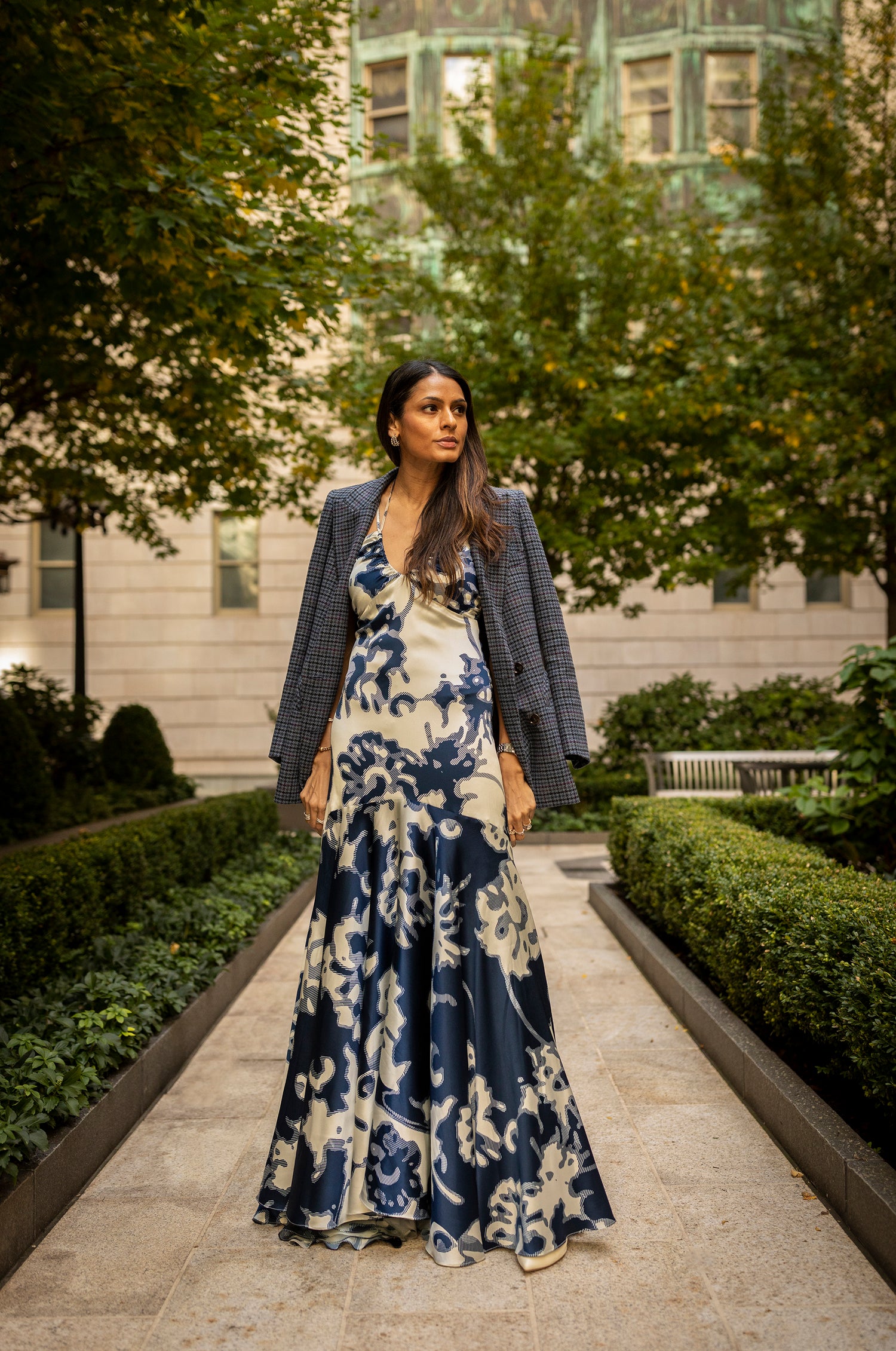 Woman in a blue and white floral gown with a plaid jacket stands on a garden path lined with bushes.