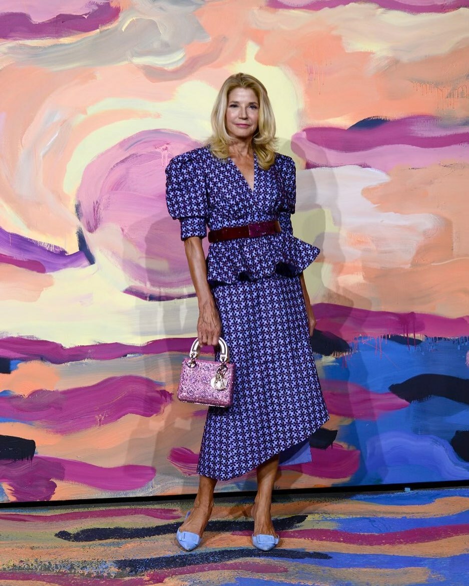 A woman in a blue patterned dress stands in front of a colorful abstract painting, holding a pink handbag.