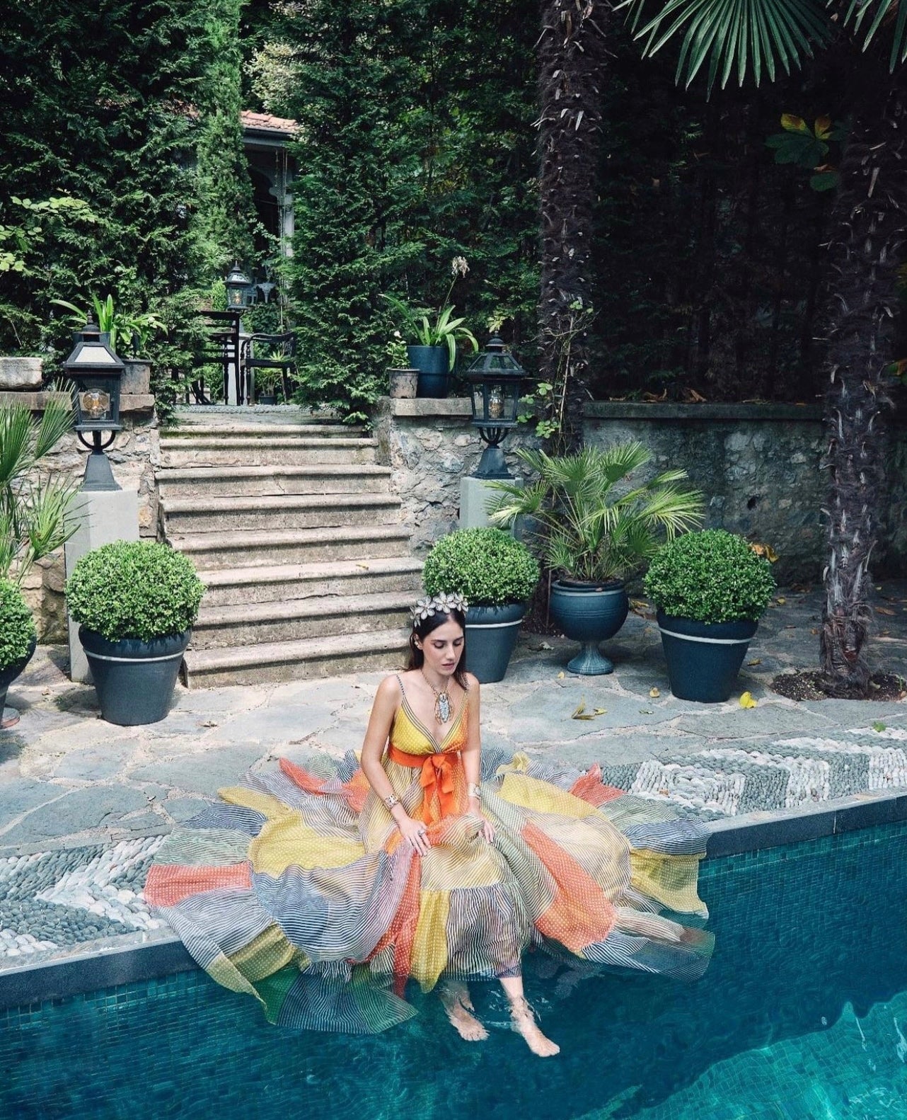 A woman in a colorful gown sits on the edge of a pool, with lush greenery and stone steps in the background.