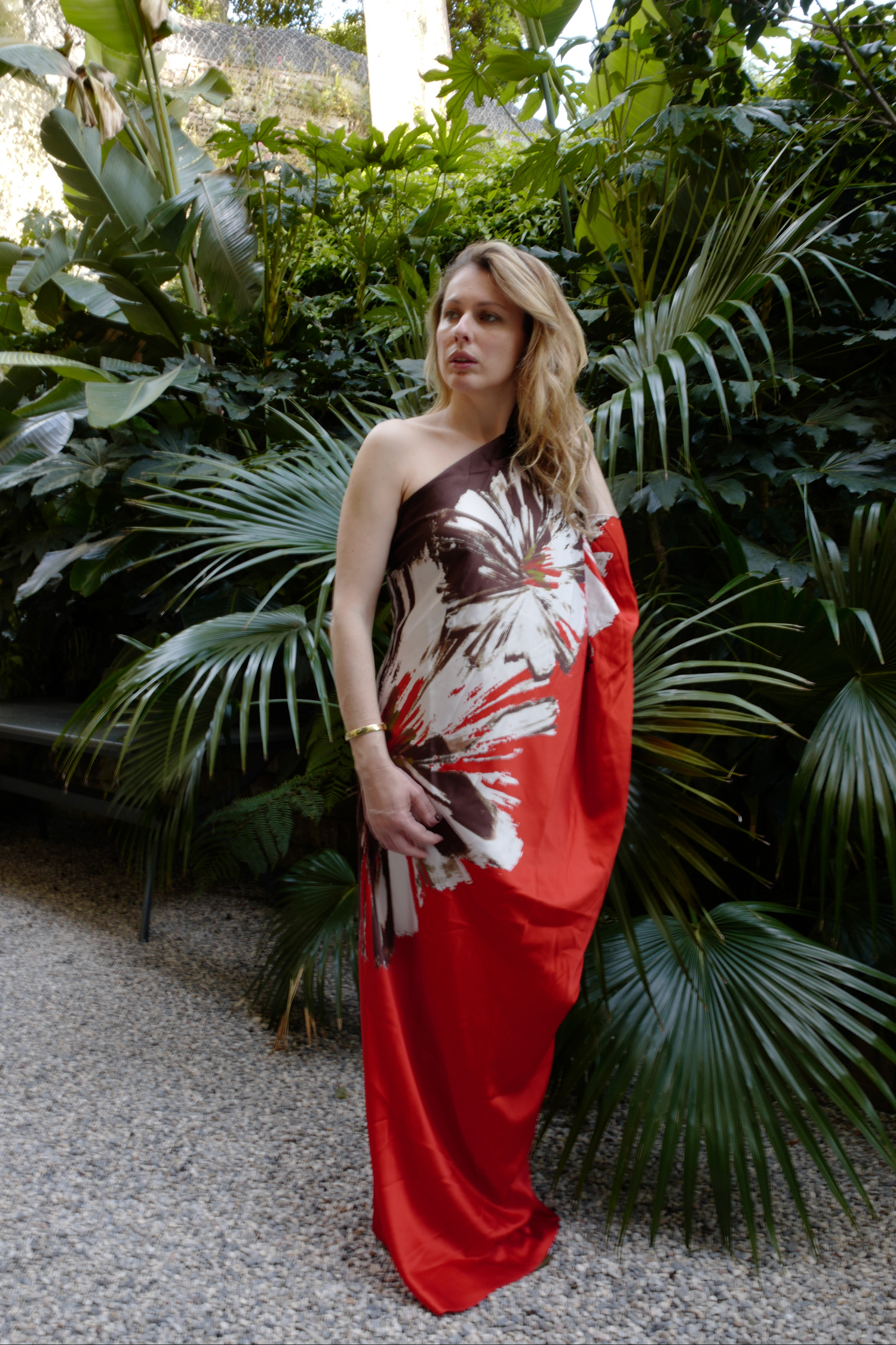 A woman in a one-shoulder red and brown floral dress stands in a garden with lush green foliage.