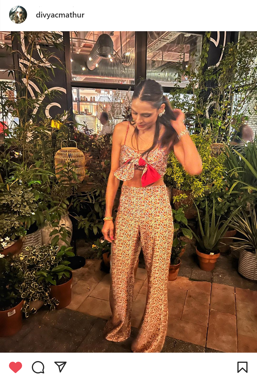 Woman in a patterned jumpsuit with a red bow stands among potted plants, looking down and adjusting her hair.