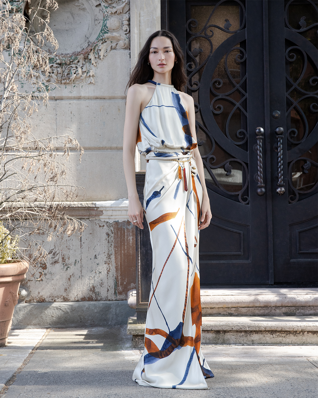 A woman in a patterned, sleeveless dress stands on a city street near an ornate, dark door.