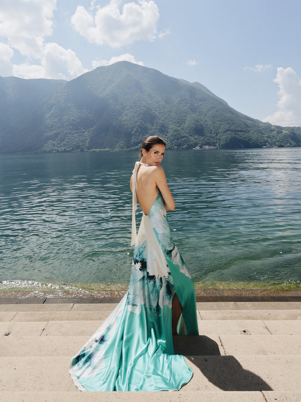 Person in a sleeveless teal gown stands on stone steps by a calm lake with mountains in the background.