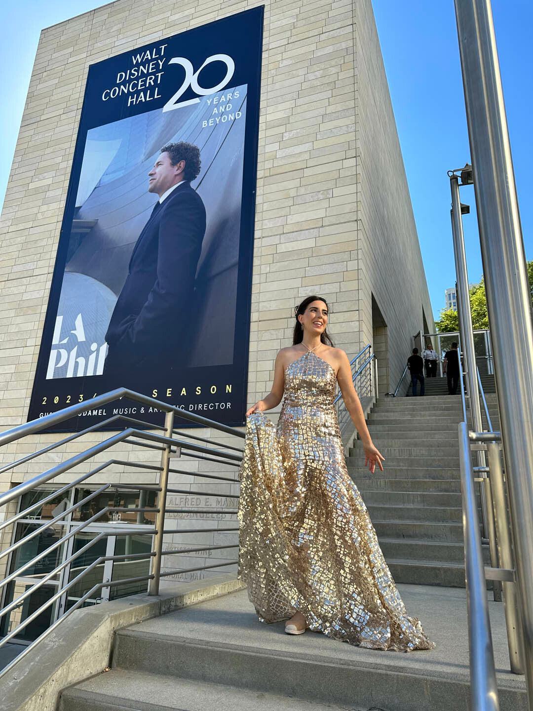 A woman in a shimmering gold dress stands on steps outside the Walt Disney Concert Hall, with a large poster on the building in the background.