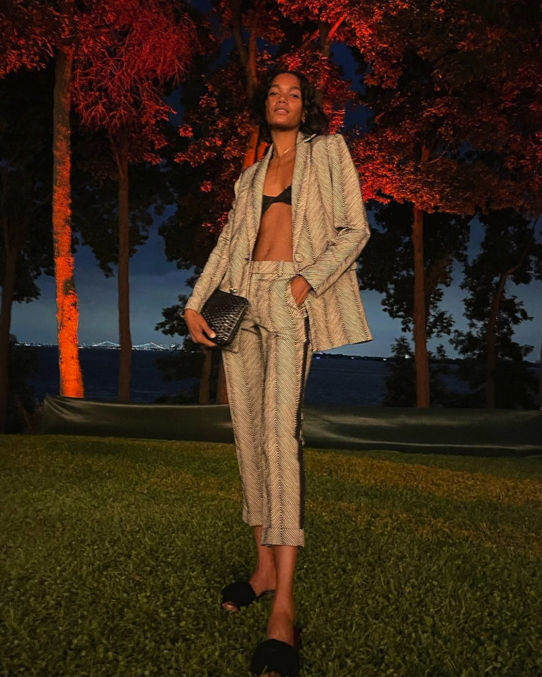 Person in a suit poses on grass at night, with red-lit trees and a dark sky in the background, holding a small bag.