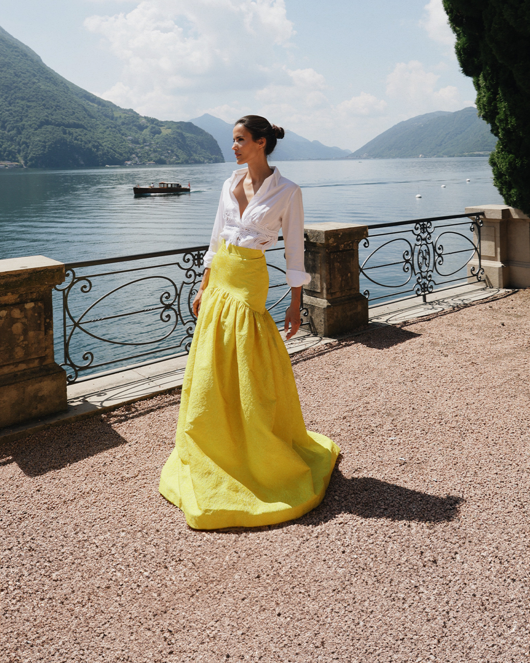 A person in a white shirt and yellow skirt stands by a lakeside railing with mountains and a boat in the background.