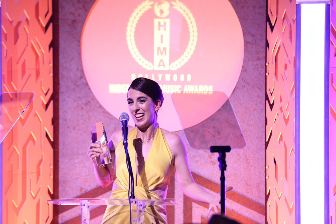 A person in a yellow dress holds an award and speaks at a podium with a microphone at the Hollywood Indie Music Awards.