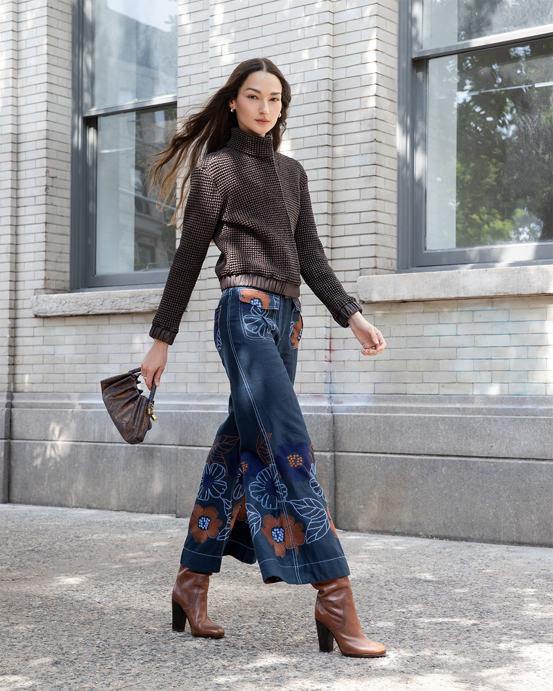 Person walking down a city street wearing a brown sweater, floral patterned pants, and brown heeled boots, holding a small purse.