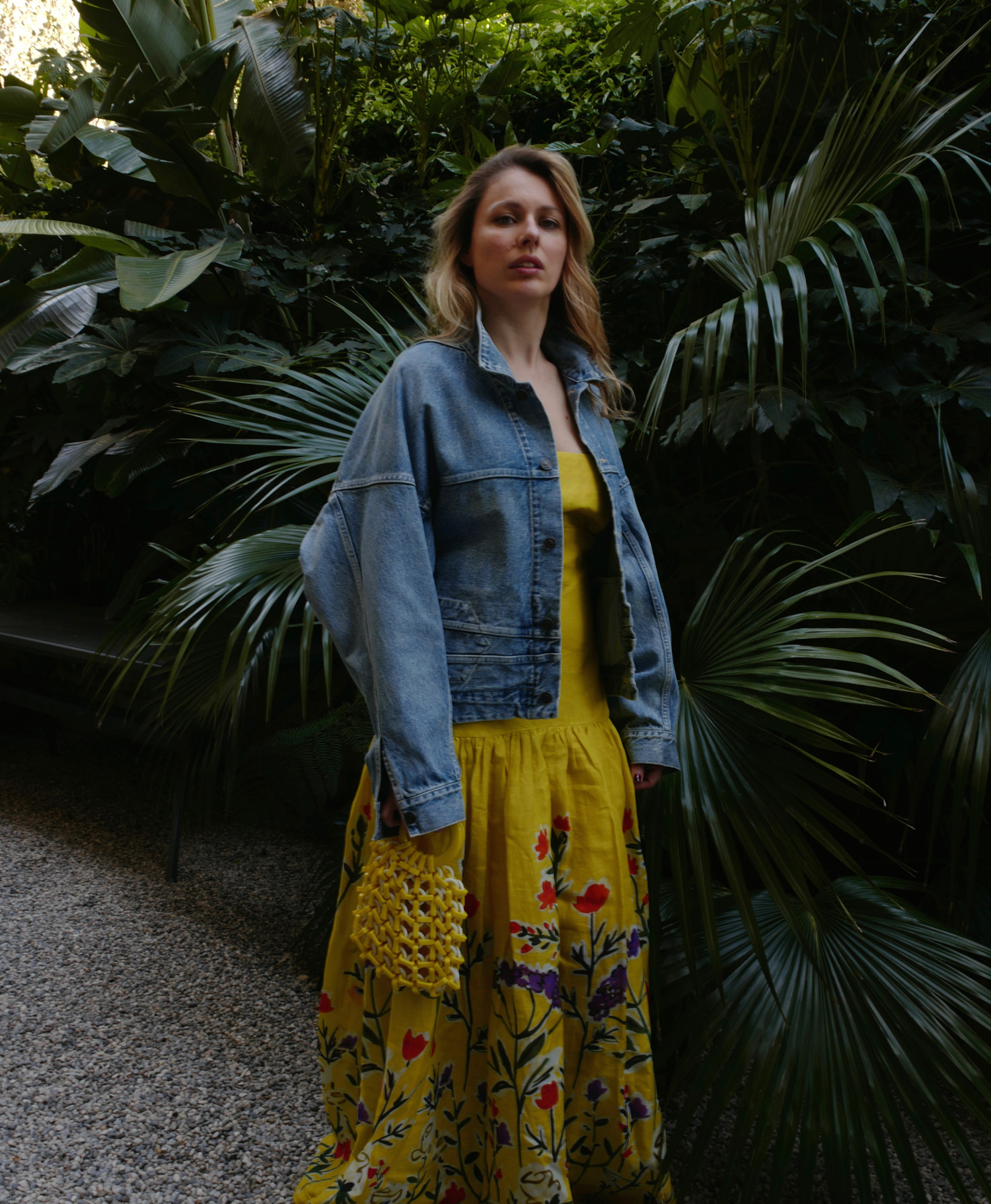 A person stands in a garden wearing a denim jacket over a yellow floral dress, holding a yellow bag.