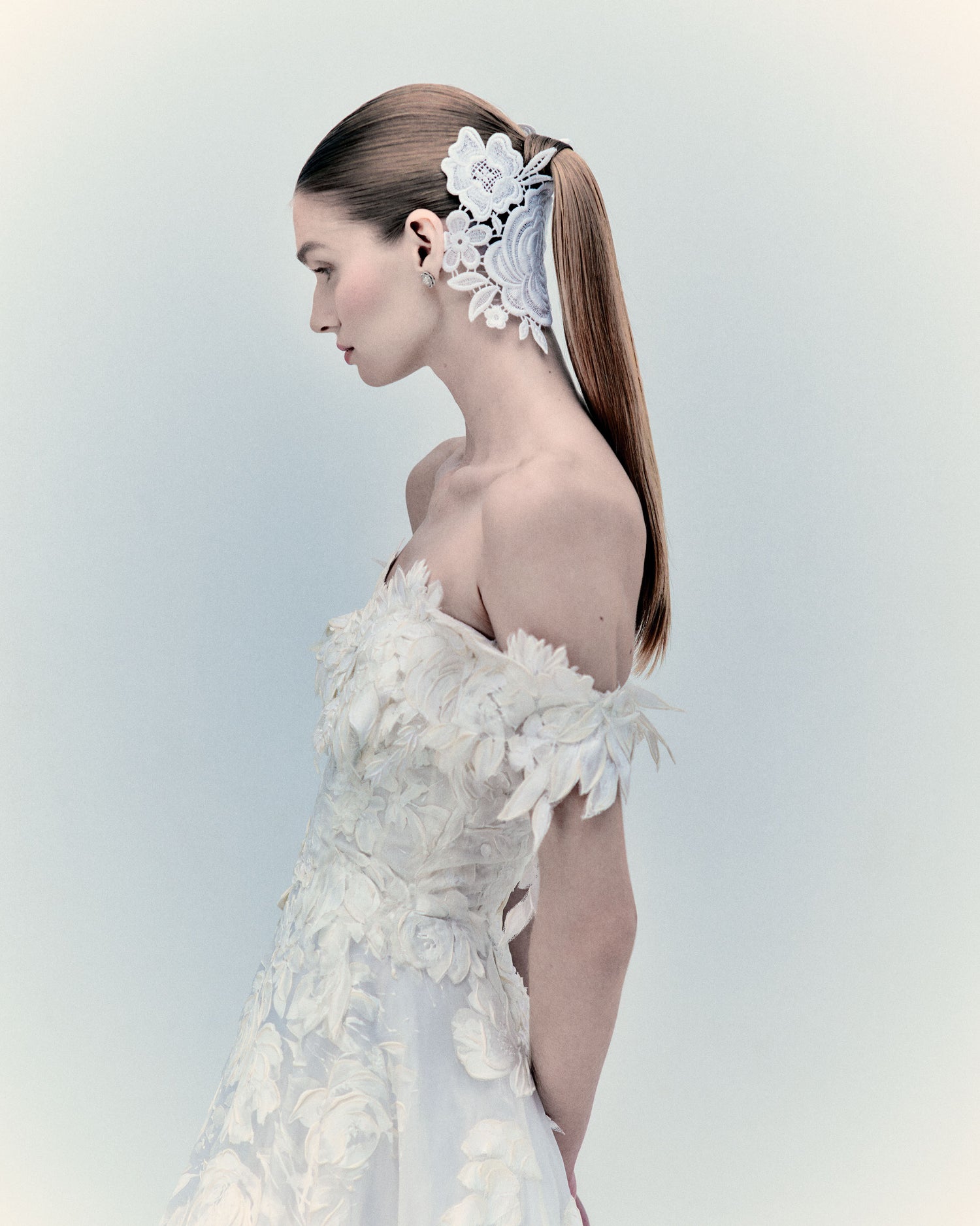 A woman in an off-shoulder, floral-textured white dress with a lace hair accessory stands in profile against a light background.