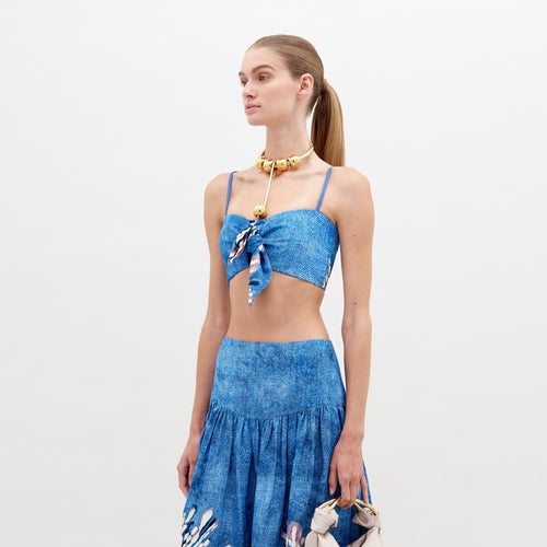 Woman with ponytail wears a blue Wotton Crop Top Aciano Flower and skirt with a large necklace. Pre-order now to ship by date, November 15th, 2024.