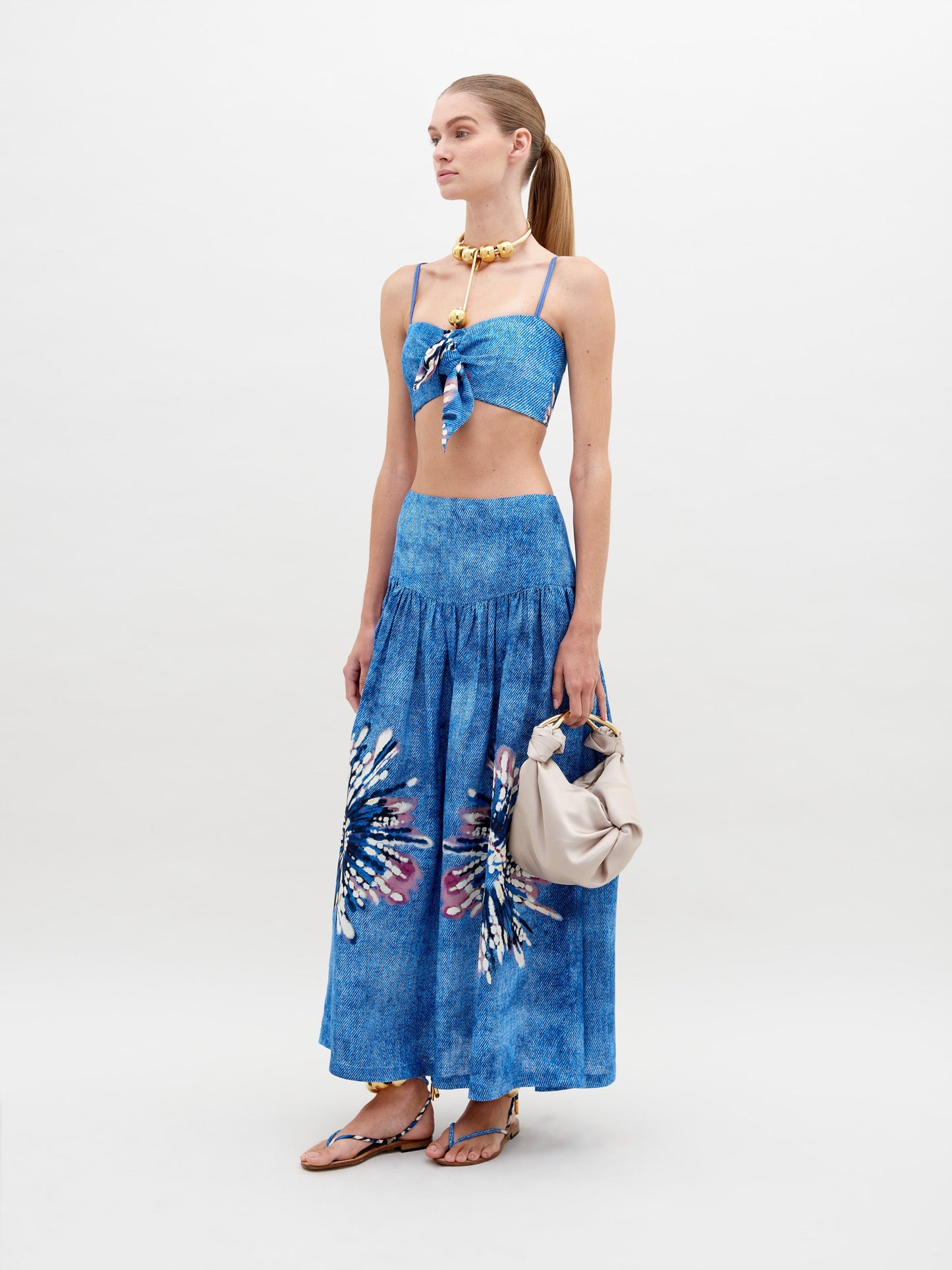 A woman wearing a blue halter top and an Abla Skirt Aciano Flower with feather patterns stands sideways, holding a light-colored, ruched handbag. With her hair in a ponytail, she epitomizes style and elegance. Pre-order now to ensure delivery by November 15th, 2024.