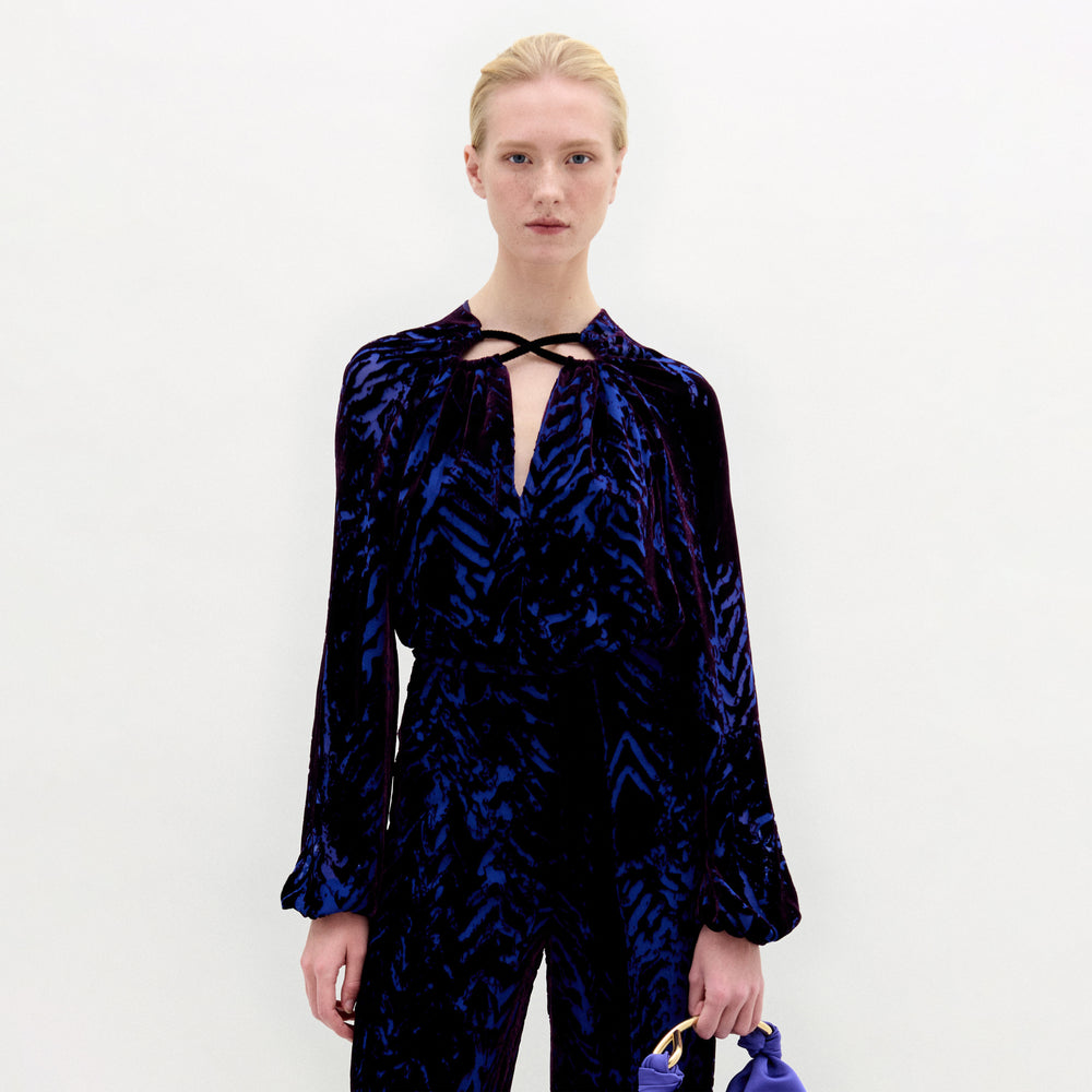 A woman wearing the Ximena Blouse in color Eggplant Velvet with a Sofia bag in color Spectrum blue on a white background