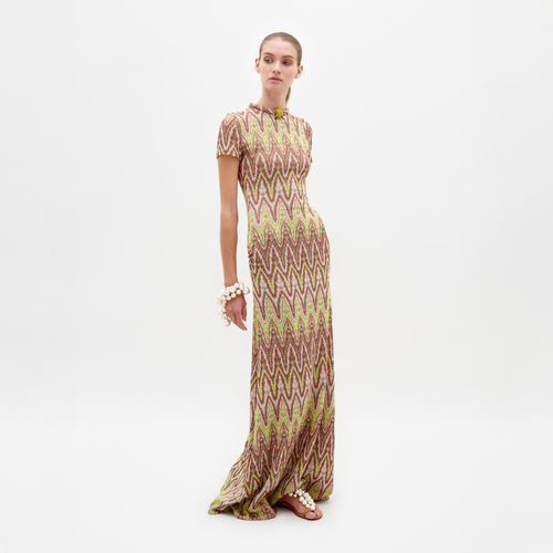 A person standing and posing in the Yara Dress Zigzag Neon, a floor-length dress with a vibrant zigzag design, adorned with large bracelet accessories and wearing flat sandals, against a white background. Pre-order now to ensure your Yara Dress Zigzag Neon ships by February 15th.