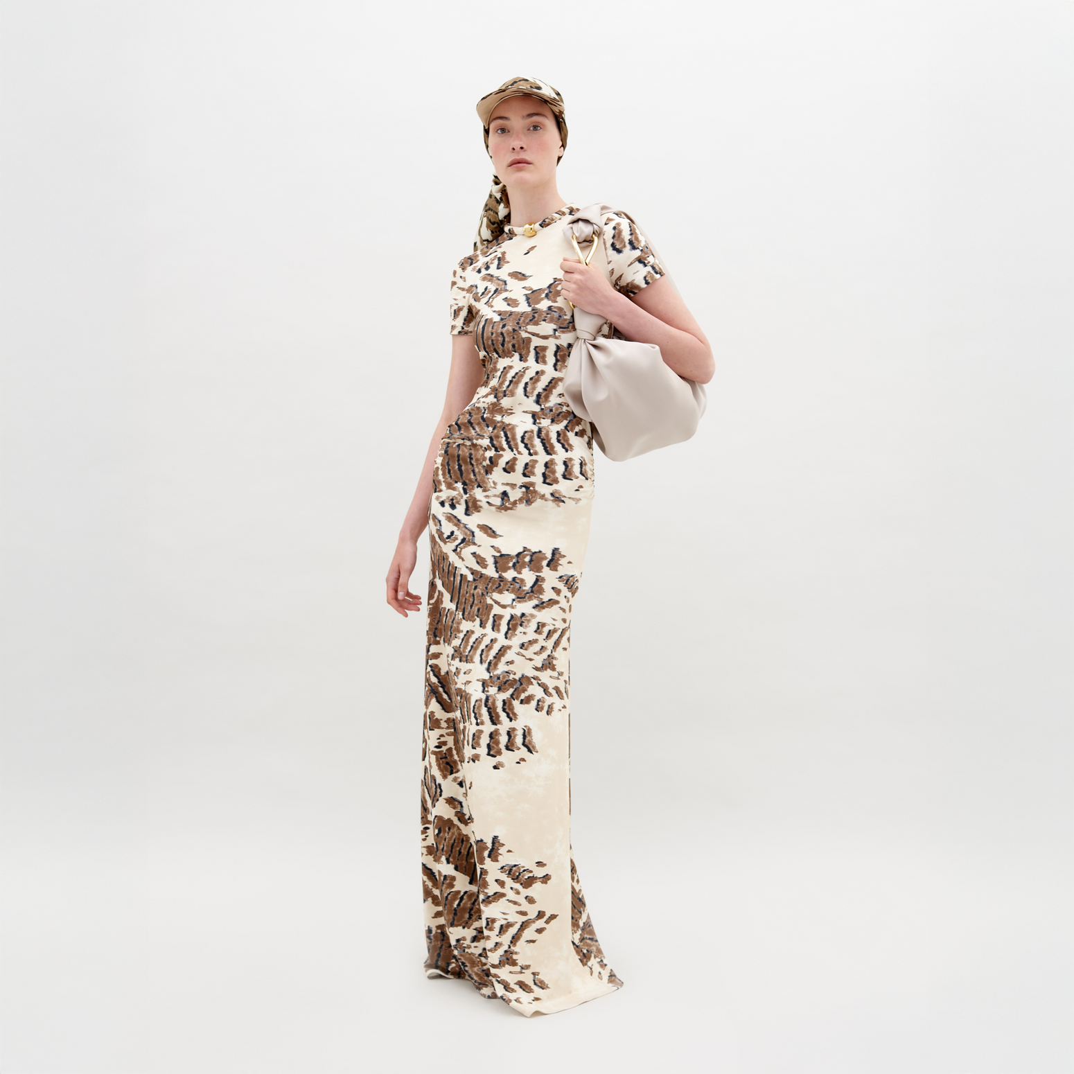 A person wearing a Yara Dress Beige Abstract Animal Print and cap with a white bag over their shoulder stands in front of a plain background, ready for pre-order by the November 15th, 2024 ship-by date.