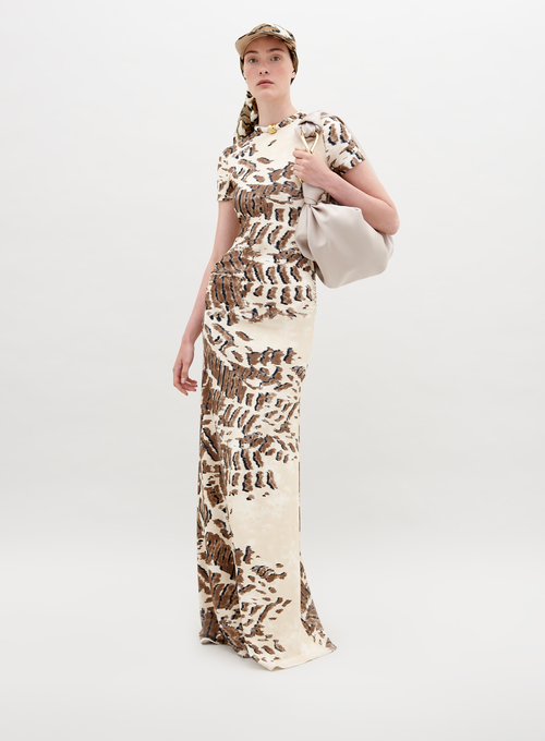 A person wearing a Yara Dress Beige Abstract Animal Print and cap with a white bag over their shoulder stands in front of a plain background, ready for pre-order by the November 15th, 2024 ship-by date.