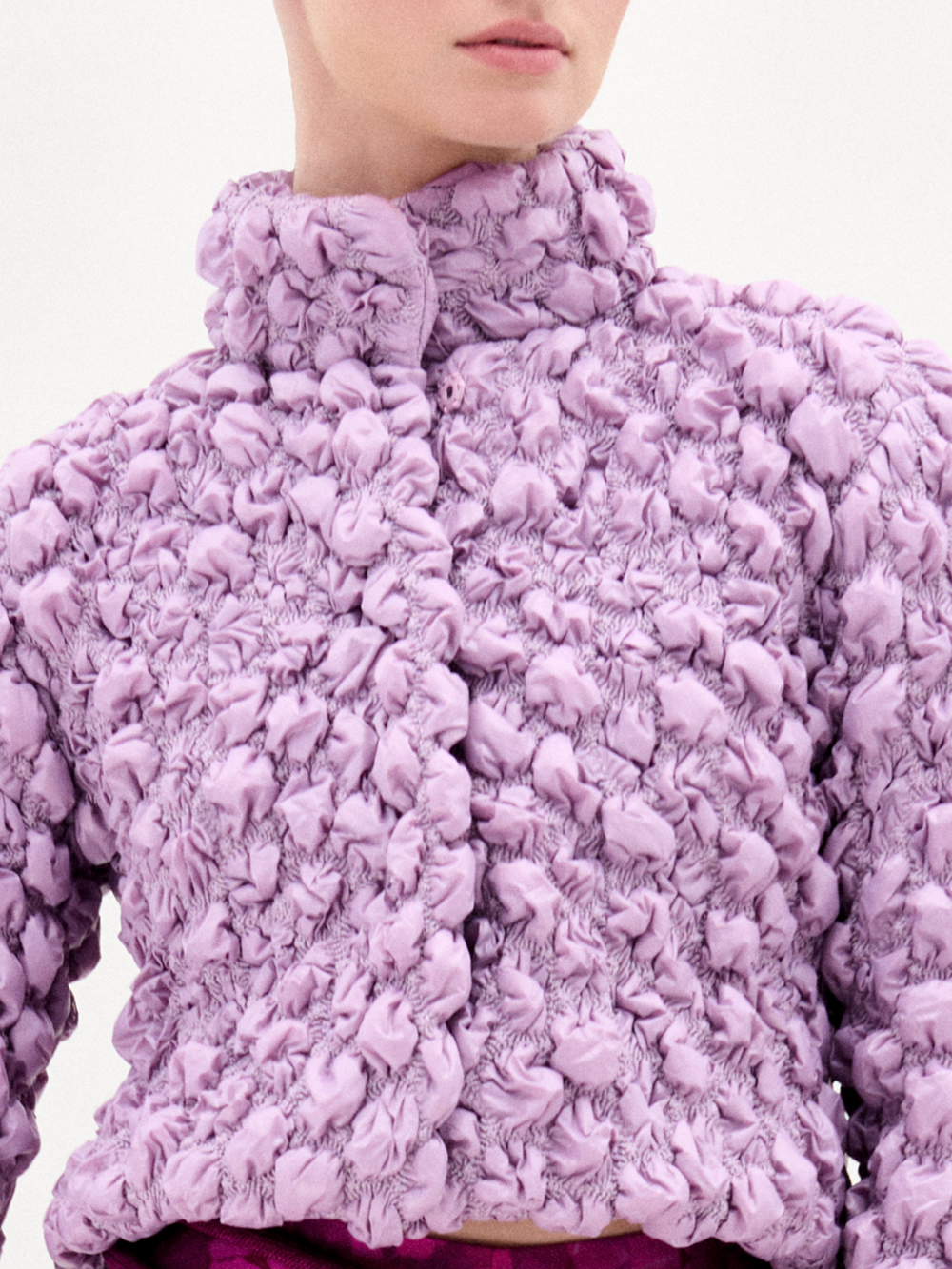 A woman poses against a white background wearing the oversized Zalta Jacket in textured lavender Italian fabric, paired with high-waisted pink patterned pants featuring a gold-buckled belt.