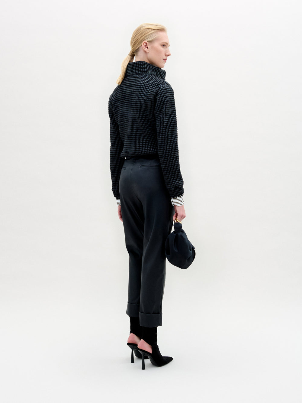 Person stands against a plain white background wearing the Moad Pant, a dark textured jacket, patterned shirt, black boots, and holding a small black handbag.