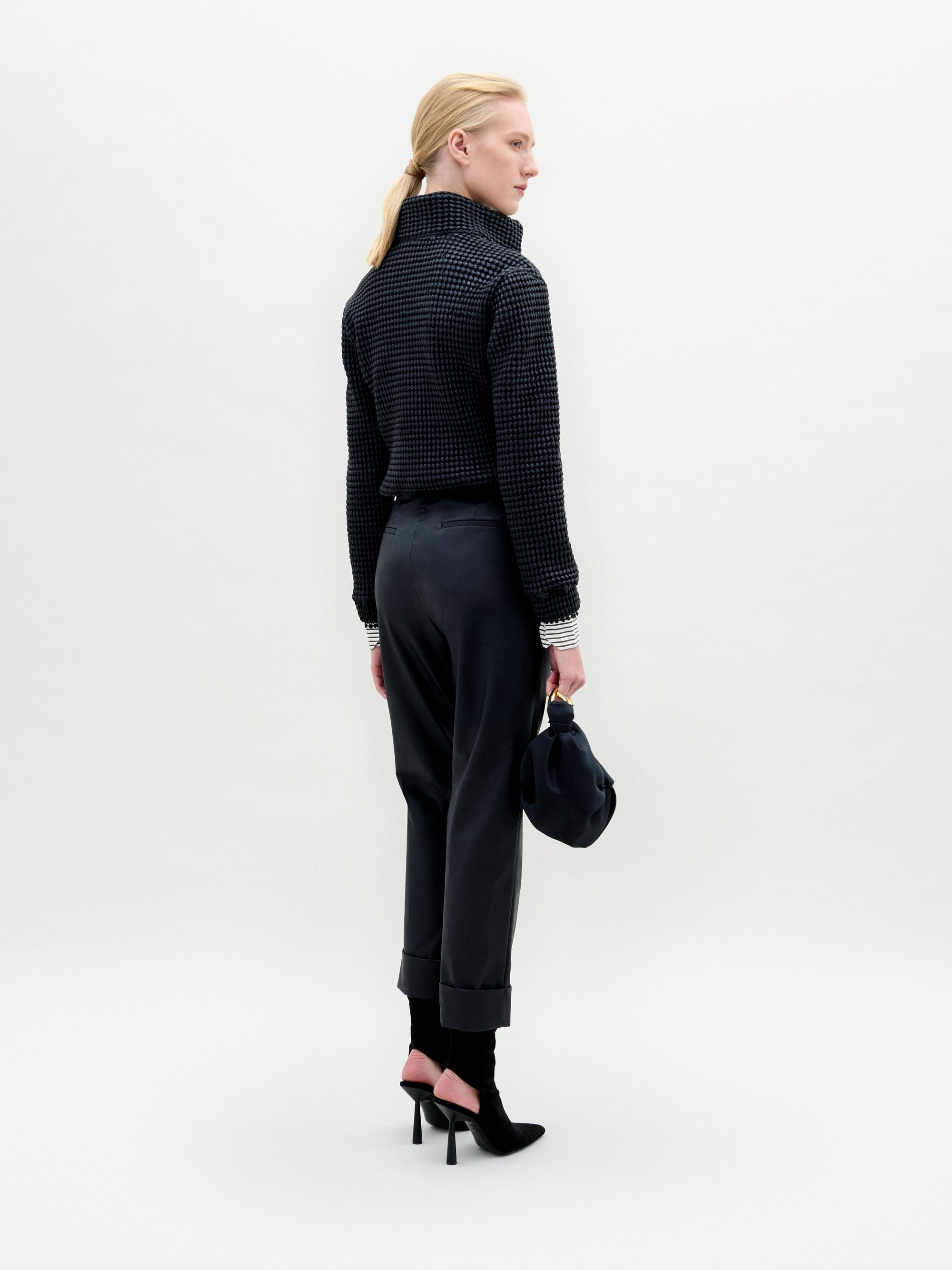Person stands against a plain white background wearing the Moad Pant, a dark textured jacket, patterned shirt, black boots, and holding a small black handbag.