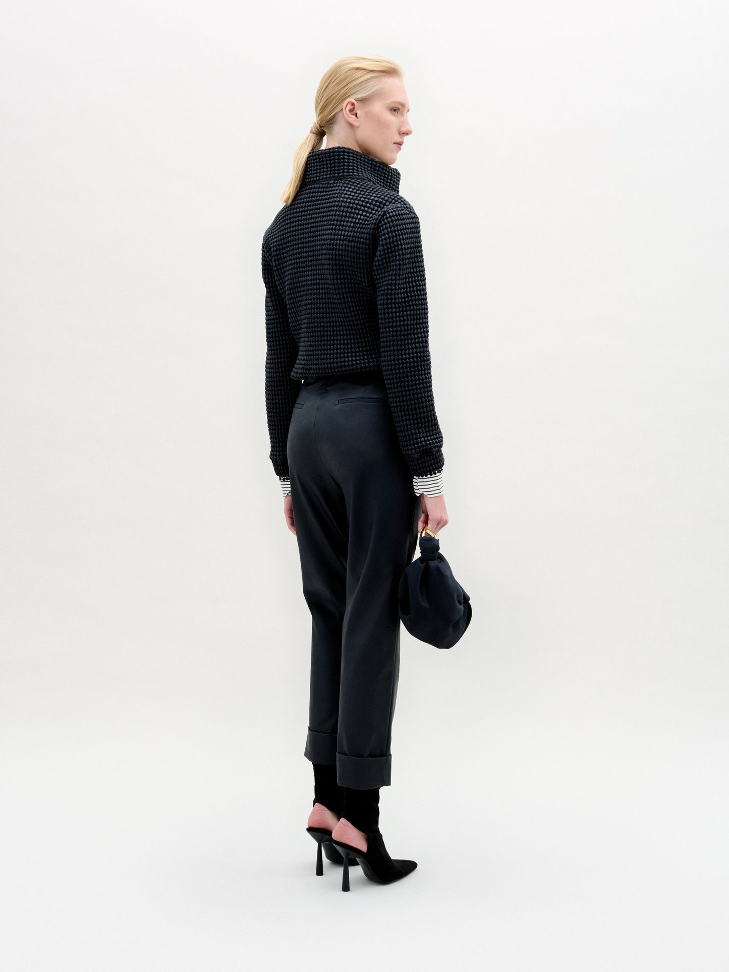 A person stands against a plain white background wearing the Zalta Jacket, a dark cropped piece crafted from Italian fabric, paired with a striped shirt, dark trousers with a gold belt, and holding a dark bag.