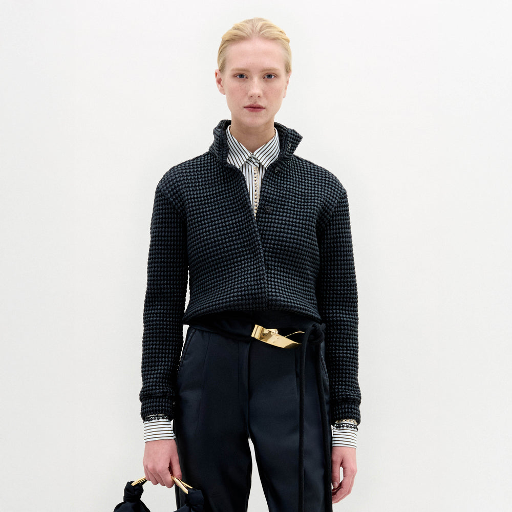 A person stands against a plain white background wearing the Zalta Jacket, a dark cropped piece crafted from Italian fabric, paired with a striped shirt, dark trousers with a gold belt, and holding a dark bag.