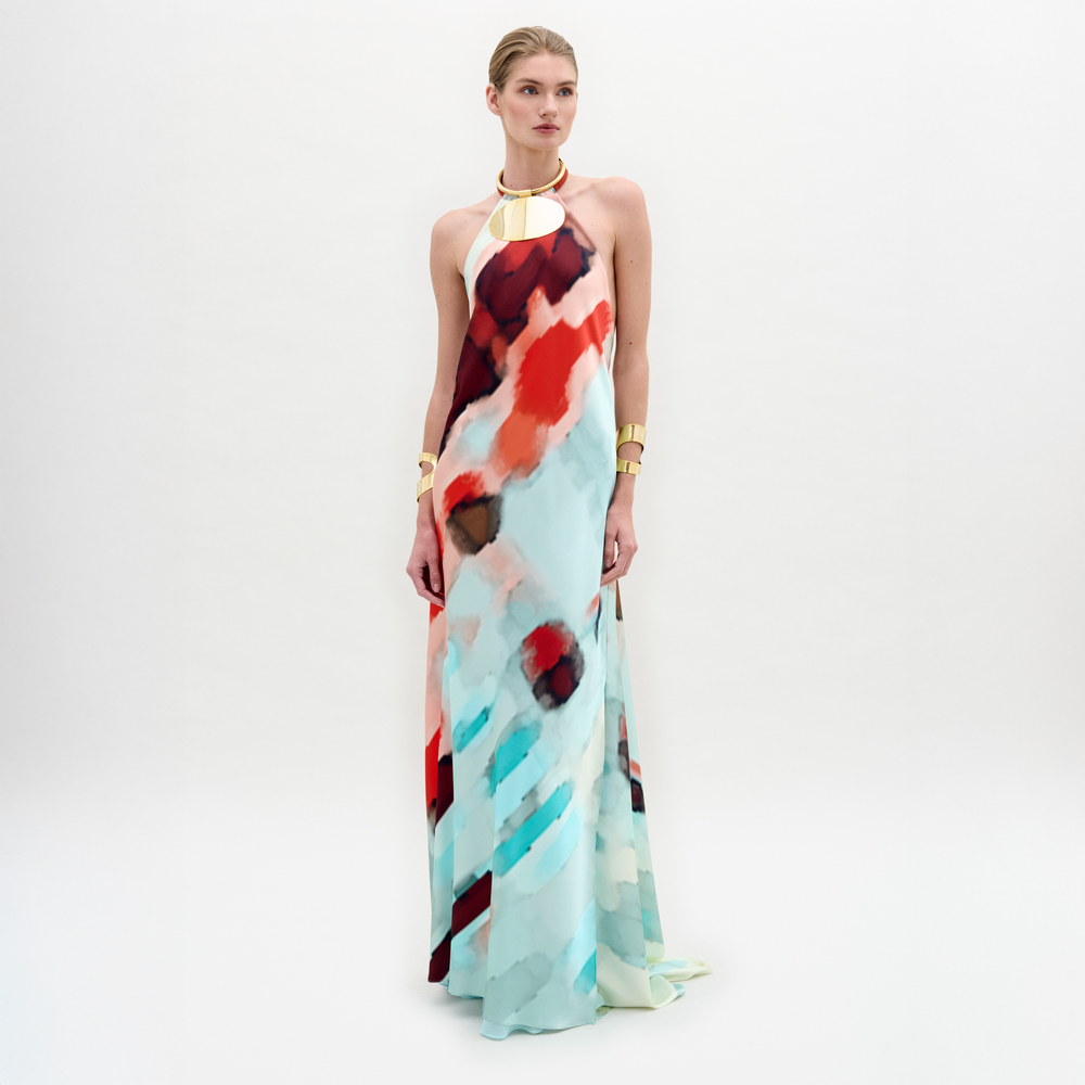 A woman poses against a white background wearing the Zeffa Dress, a sleeveless, floor-length silk maxi with an abstract red, aqua, and white pattern, paired with gold jewelry.