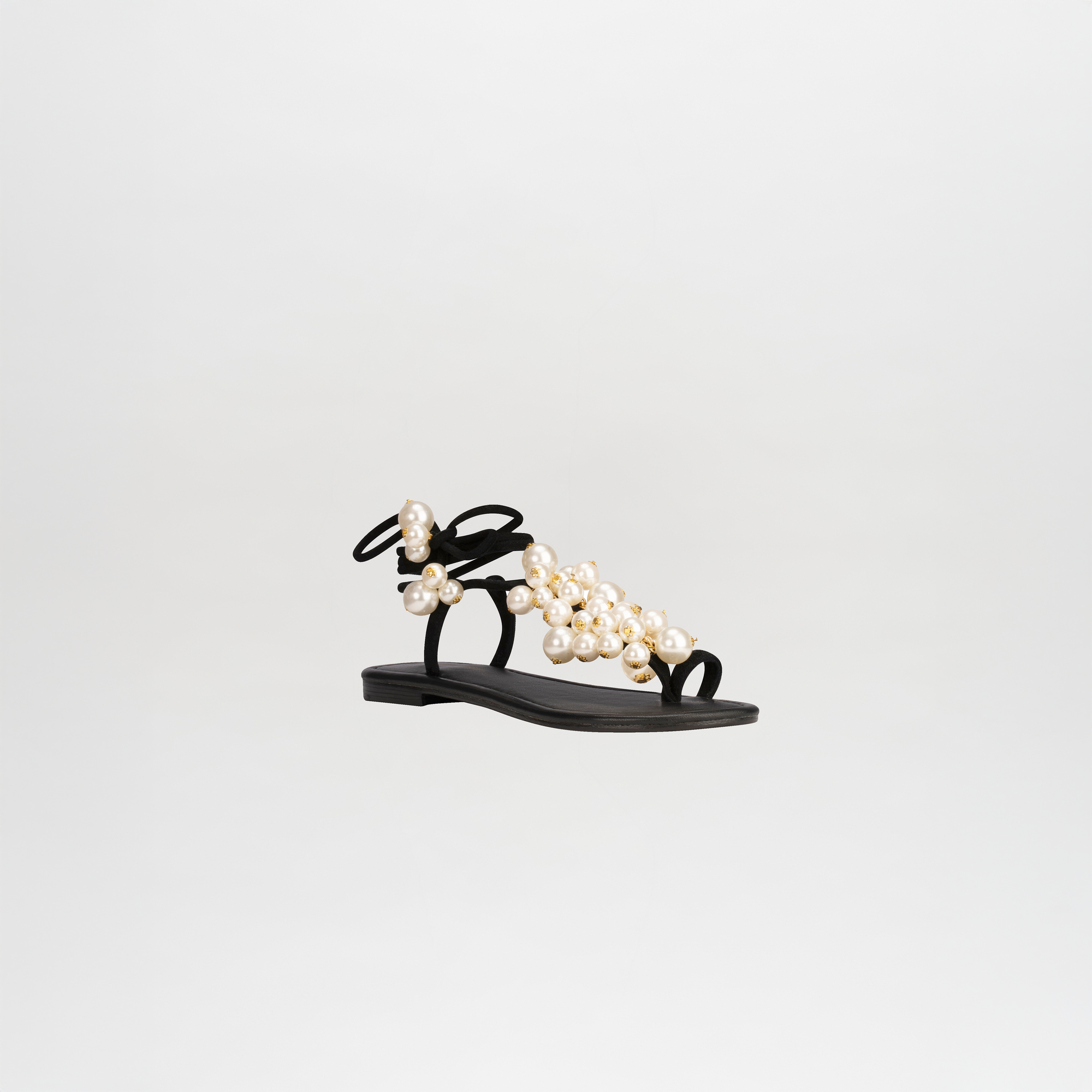 The Zaira Sandals are chic black leather footwear featuring pearl-embellished straps, a flat sole, and elegant wrap-around ankle straps, all elegantly presented against a white background.