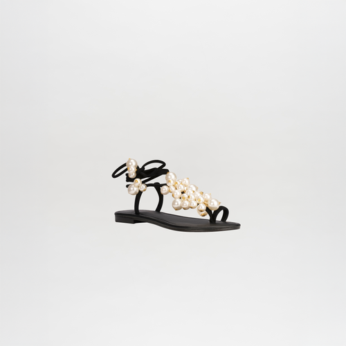 The Zaira Sandals are chic black leather footwear featuring pearl-embellished straps, a flat sole, and elegant wrap-around ankle straps, all elegantly presented against a white background.