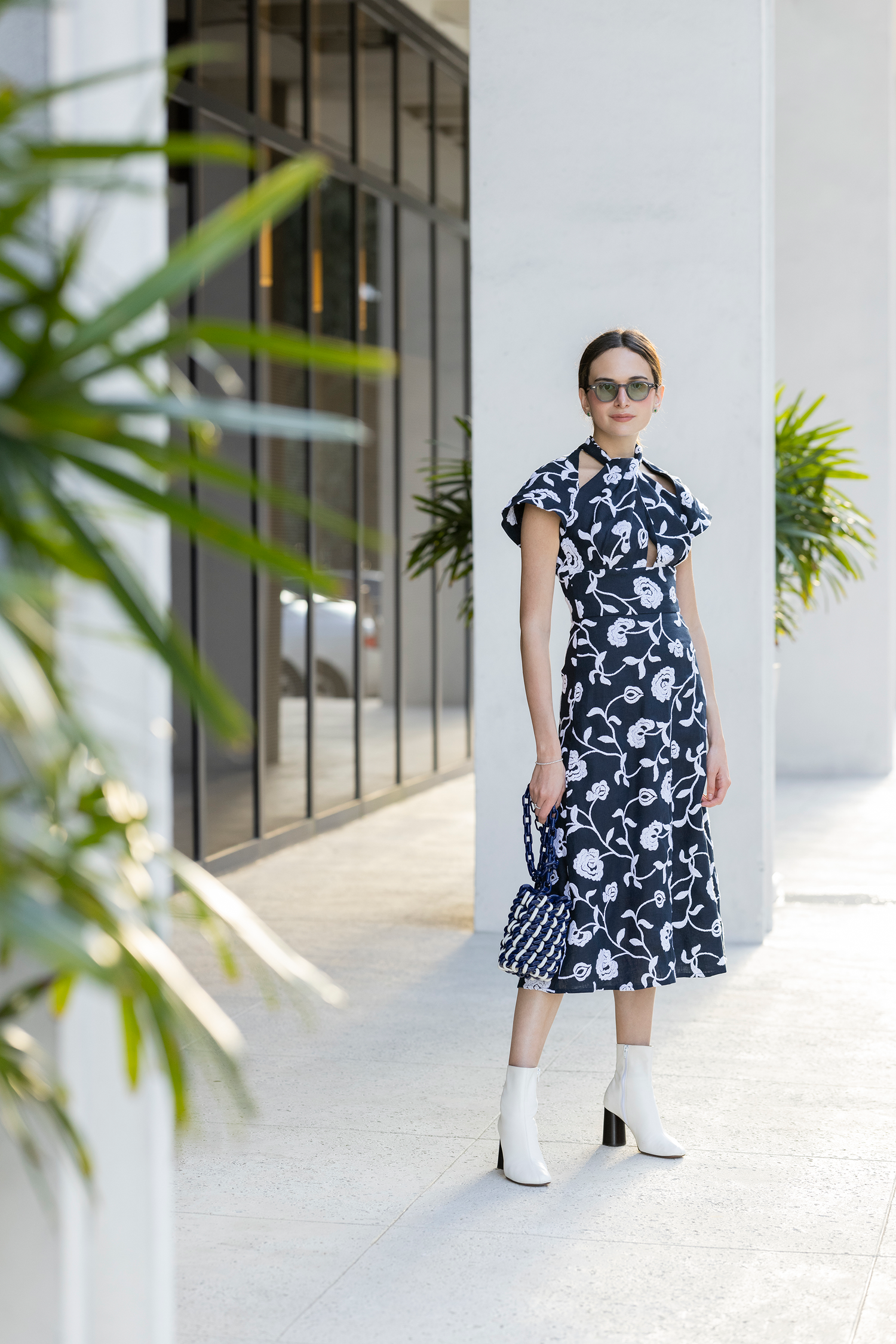 A person in a black and white floral dress and sunglasses stands on a sidewalk near a building with large windows. 