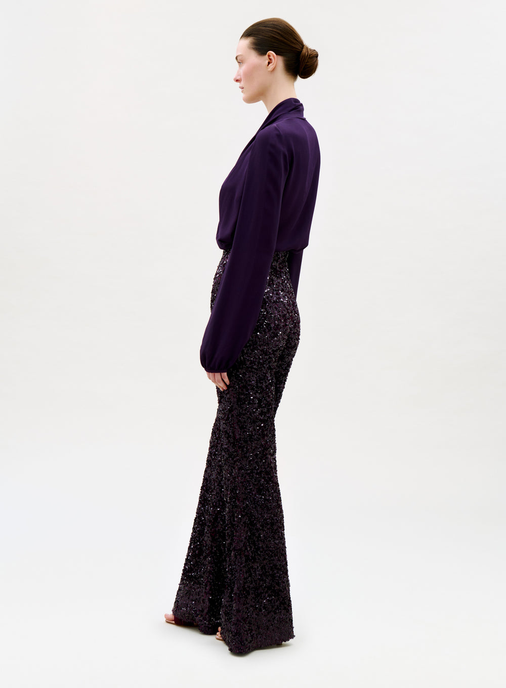 A person stands in profile against a plain background, wearing a long-sleeved, deep purple blouse and the Avellino Pant—high-waisted, wide-leg sequined flared pants in a matching shade—with hair pulled back in a neat bun.