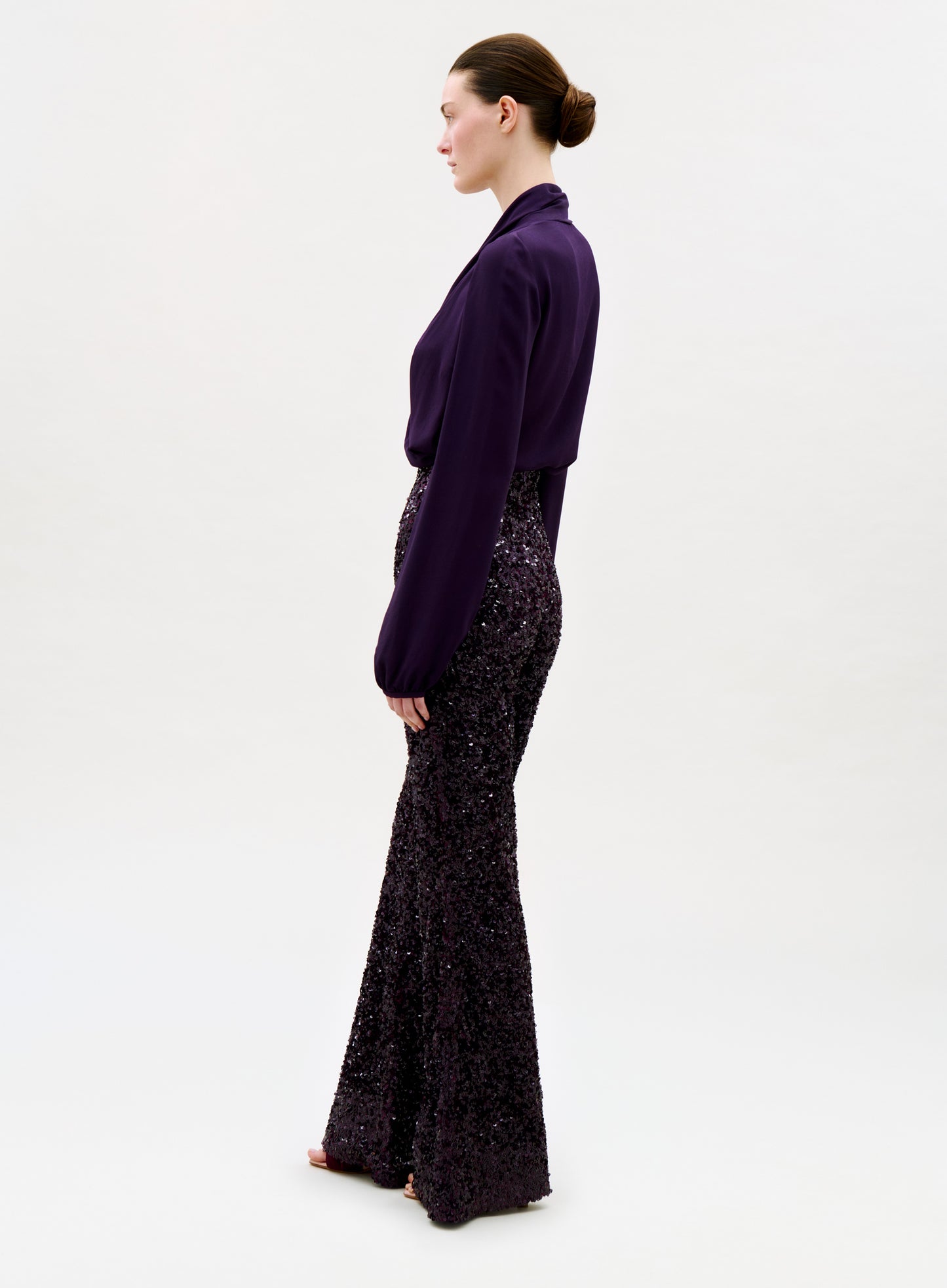 A person stands in profile against a plain background, wearing a long-sleeved, deep purple blouse and the Avellino Pant—high-waisted, wide-leg sequined flared pants in a matching shade—with hair pulled back in a neat bun.