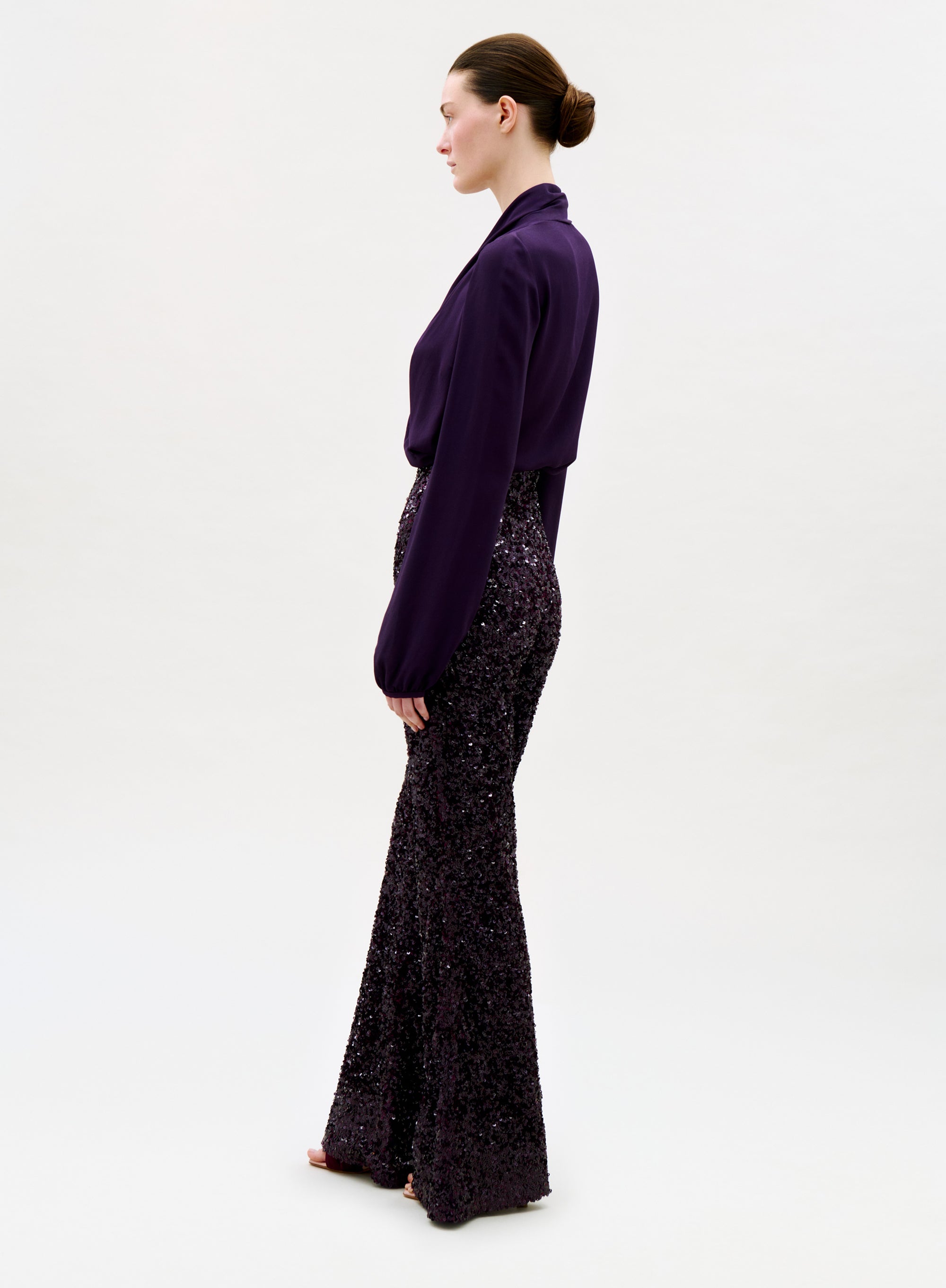 A person stands in profile against a plain background, wearing a long-sleeved, deep purple blouse and the Avellino Pant—high-waisted, wide-leg sequined flared pants in a matching shade—with hair pulled back in a neat bun.