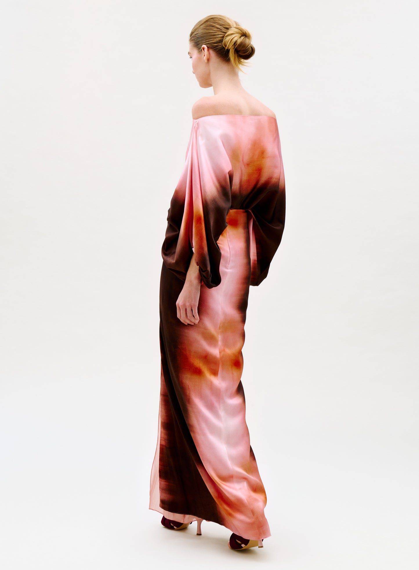 A person with blonde hair in a low bun models the Keiry Skirt, a high-waisted, off-the-shoulder maxi gown with long sleeves and a pink, brown, and orange watercolor pattern, crafted from luxurious Italian fabric against a plain light background.
