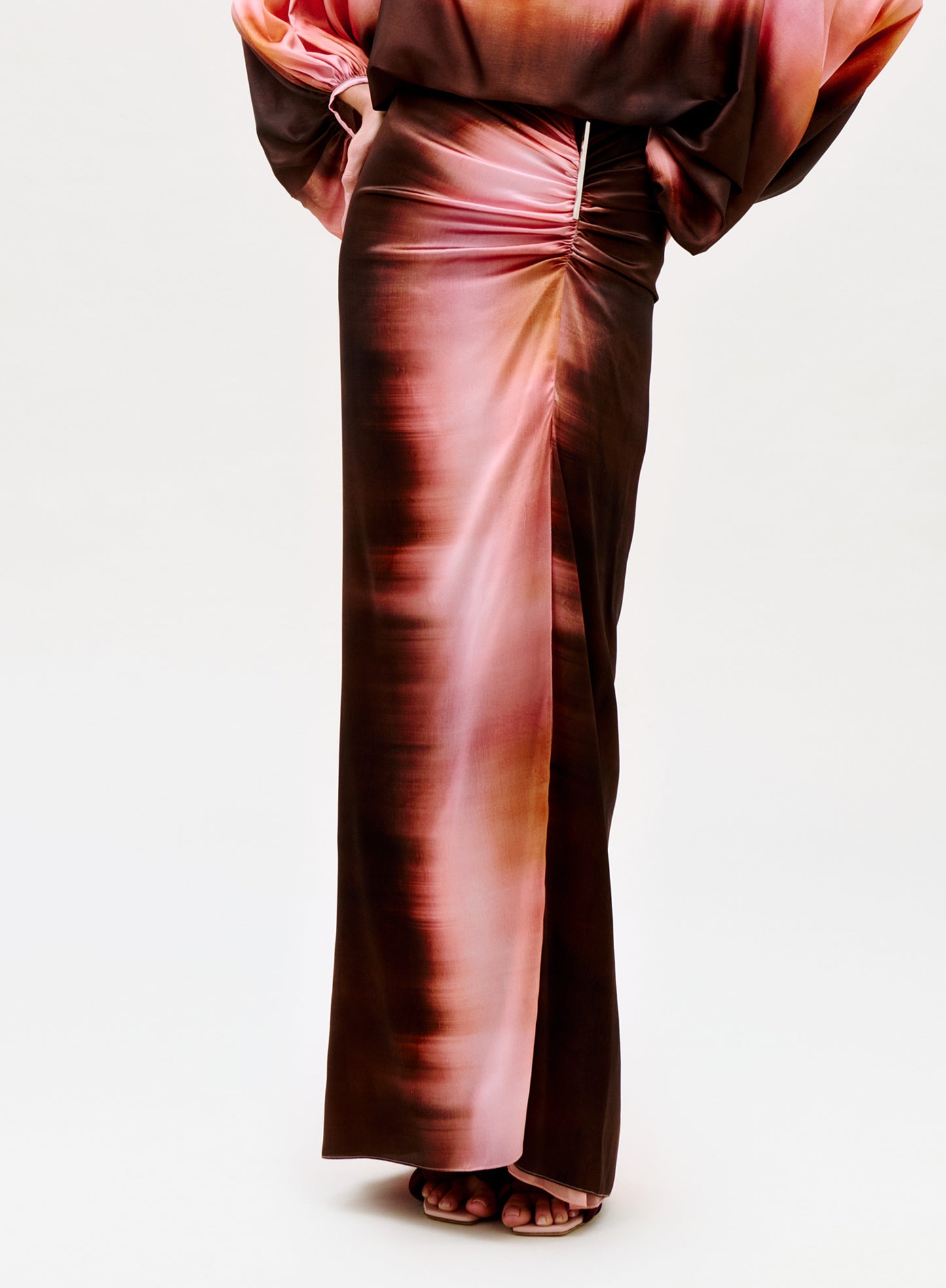 A person wearing the Keiry Skirt, a high-waisted silky maxi in pink and brown with a sheen, ruched waist, high slit, and matching loose-sleeved top. Feet are shown in open-toe shoes.
