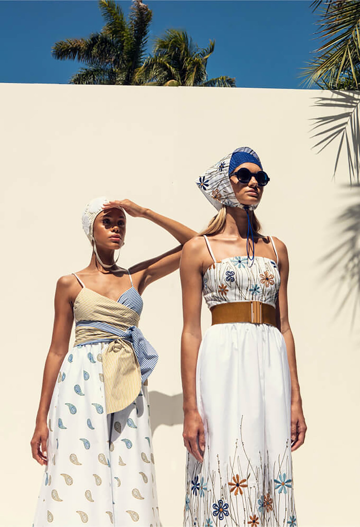 Two women in patterned summer dresses and headscarves stand outdoors against a white wall with palm trees in the background.